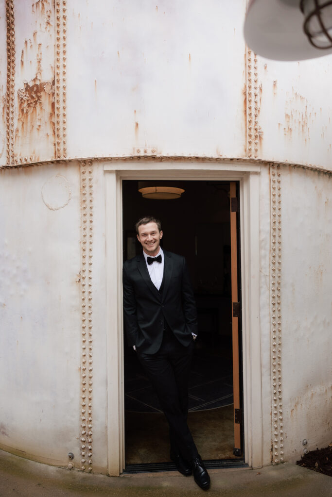 Groom in a black tuxedo smiling in the doorway of a vintage metal building at Halter Ranch