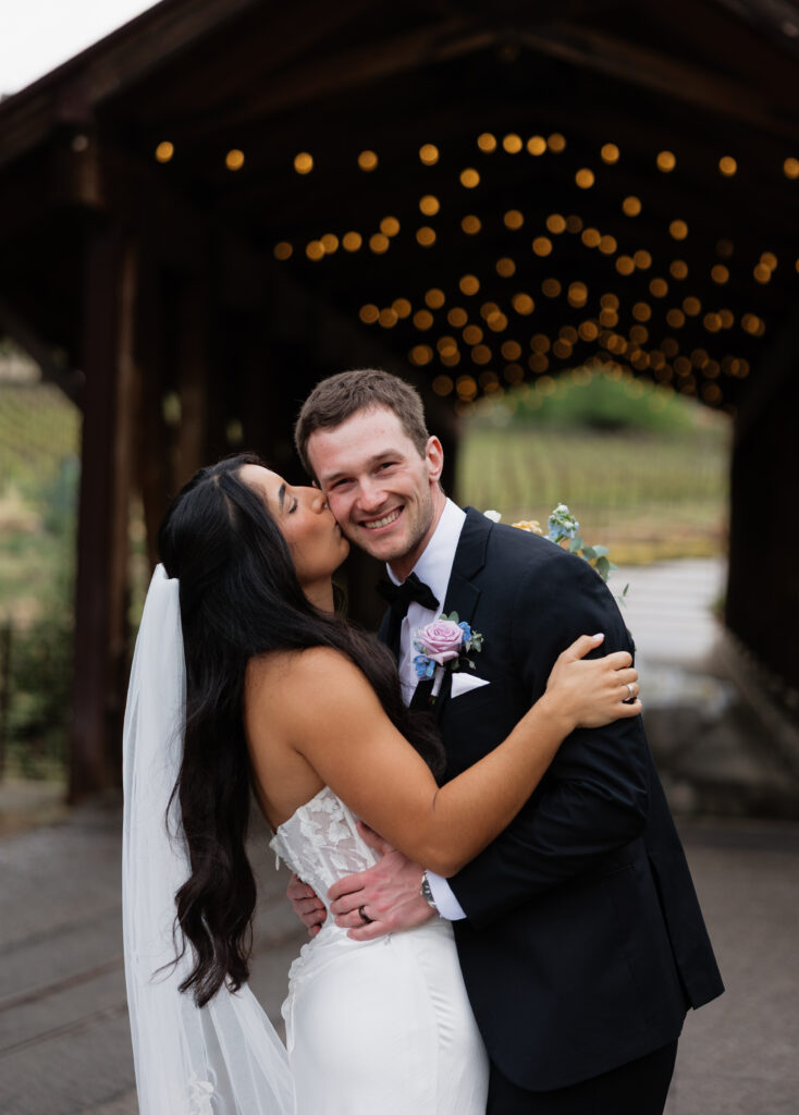 Bride kissing the groom beneath warm string lights at the covered bridge